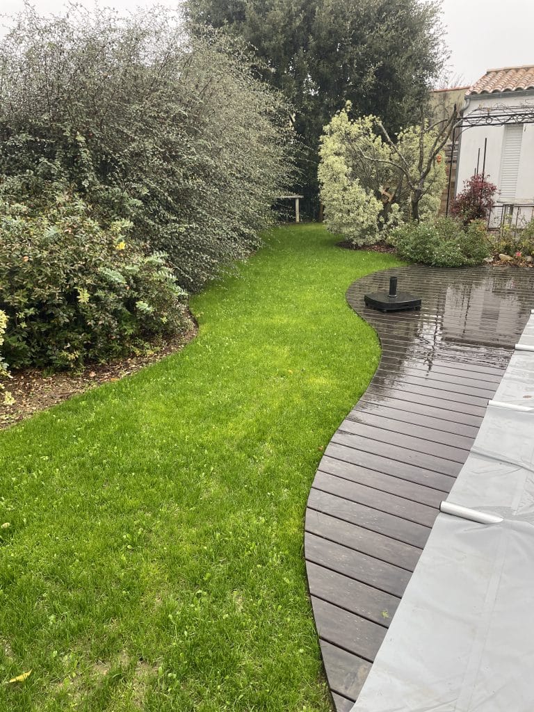 Jardin paysager avec pelouse tonte naturelle aux formes courbes , bordée d’une terrasse en bois et de massifs végétalisés.