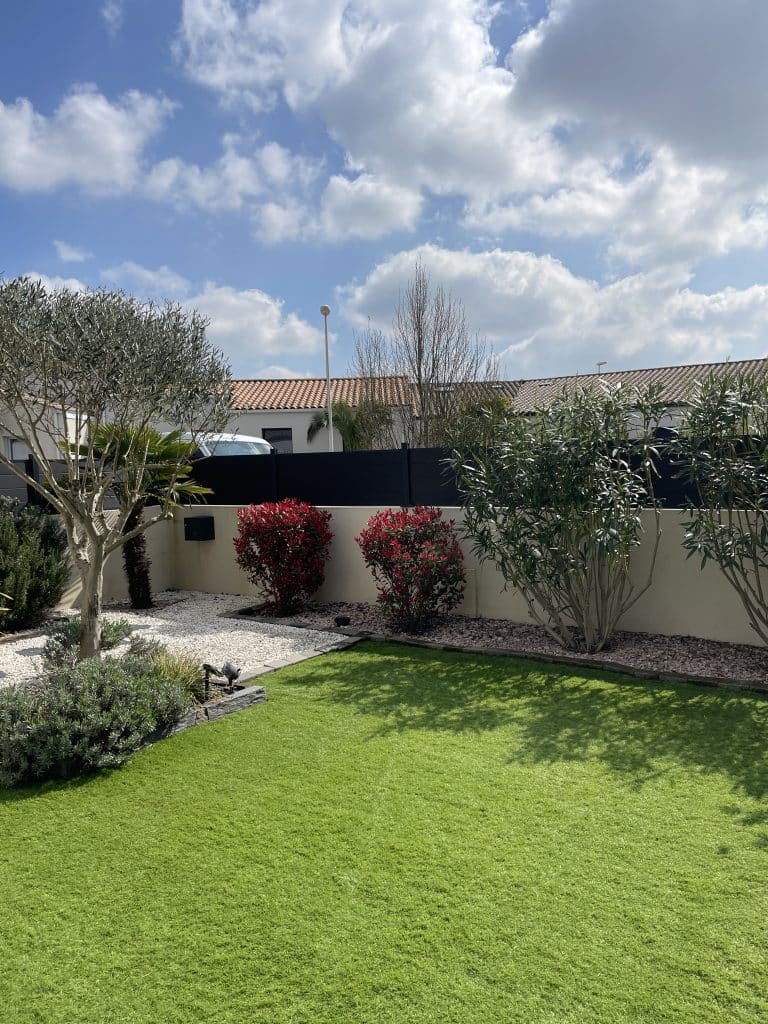 Aménagement de jardin avec pelouse verte, plantations décoratives et arbustes le long d’un mur de clôture.