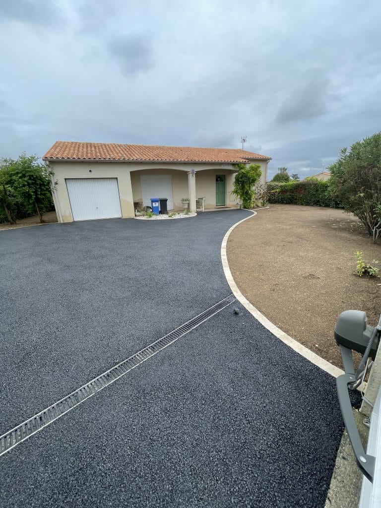 Allée d’accès en enrobé noir avec bordure claire, menant à une maison individuelle avec garage.