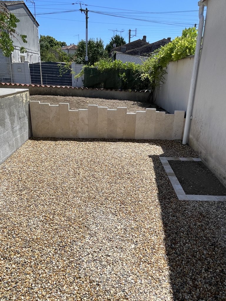 Aménagement extérieur en gravier avec bordures en pierre et muret de soutènement, espace propre et structuré le long d’un mur.