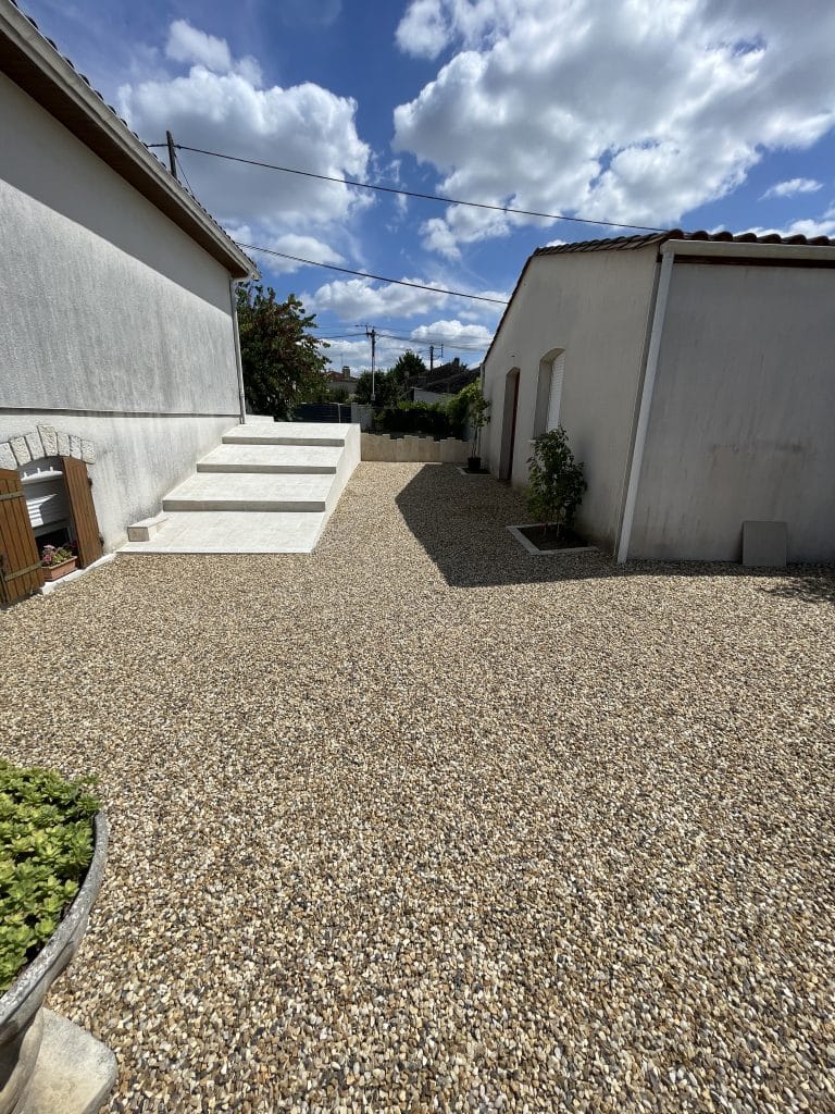 Cour extérieure aménagée en gravier décoratif avec escalier en béton, entre une maison et une dépendance, finition moderne et drainante.