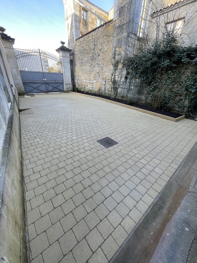Cour intérieure pavée en pavés béton clairs, avec caniveau central et bordures paysagères le long d’un mur en pierre, finition soignée et durable.