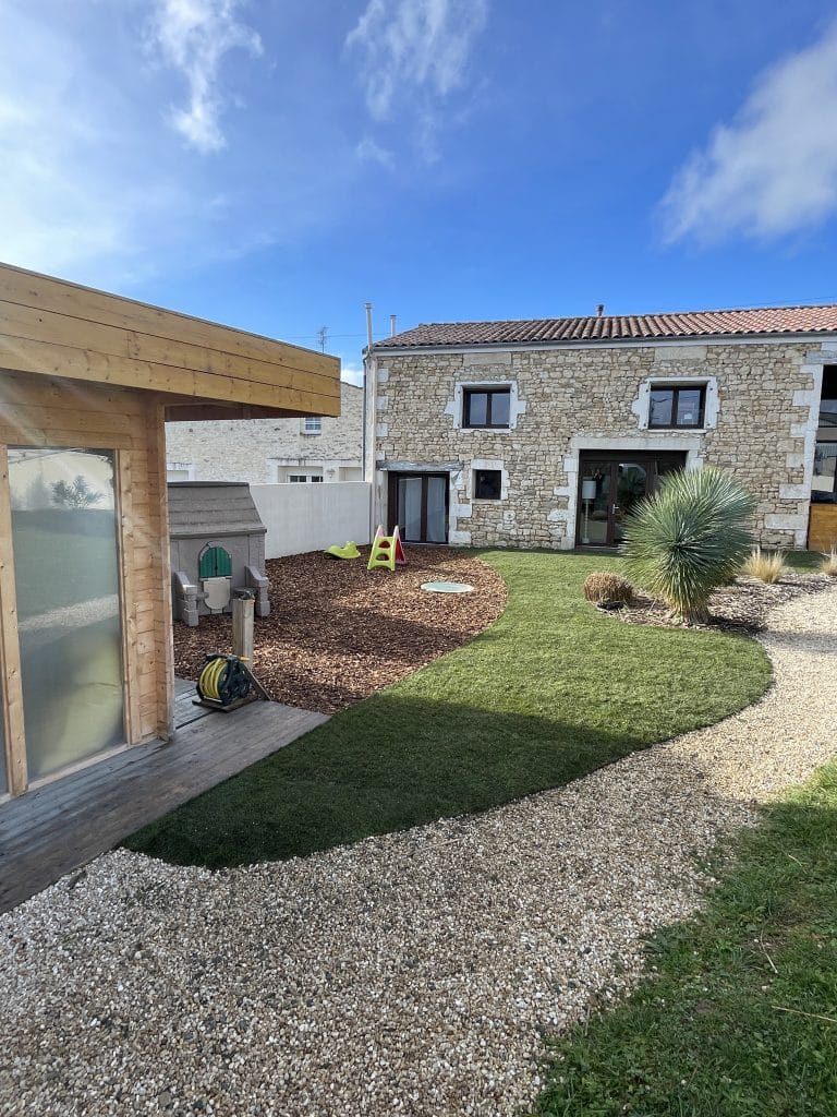 Jardin aménagé avec pelouse aux formes courbes, massif en copeaux de bois et graviers, espace ludique pour enfants et plantations décoratives autour d’une maison en pierre.